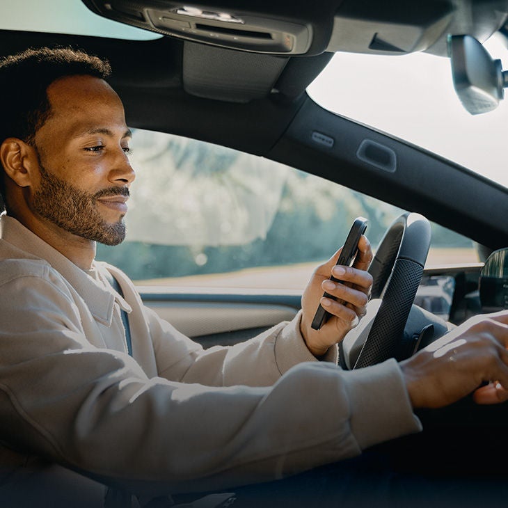 Mann sitzt im Auto mit Handy in der Hand und bedient gleichzeitig das Infotainmentsystem.